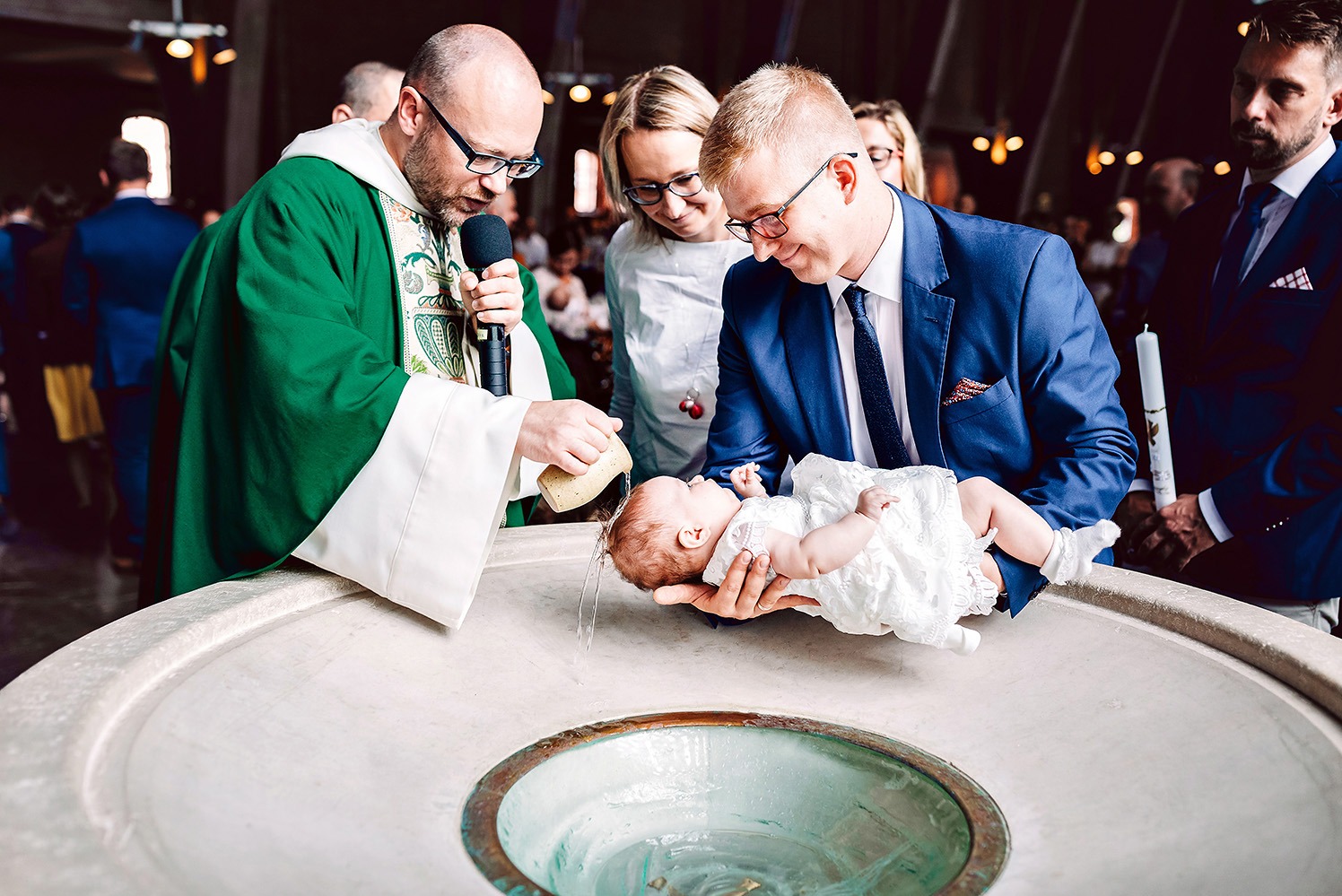 Ksiądz polewa wodą główkę niemowlęcia trzymanego przez ojca nad kamienną chrzcielnicą. W tle matka i goście. Uroczystość chrztu w kościele.