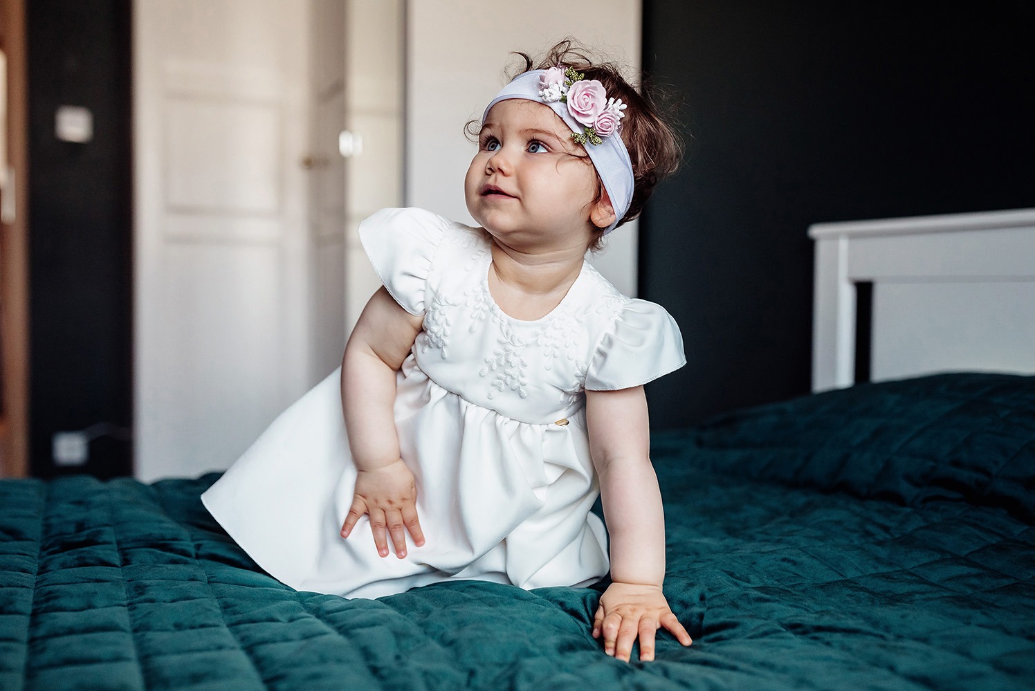 Urocza dziewczynka w białej sukience i opasce z kwiatami siedzi na łóżku z zieloną narzutą, spoglądając w górę. Delikatne światło podkreśla jej dziecięcą twarz.