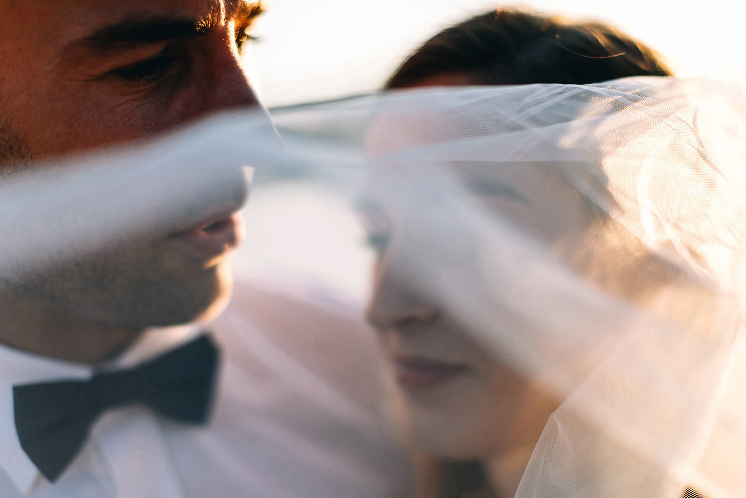 Młoda para w zbliżeniu, częściowo zasłonięta woalką. Pan młody w muszce, delikatne światło podkreśla rysy twarzy. Romantyczna atmosfera.