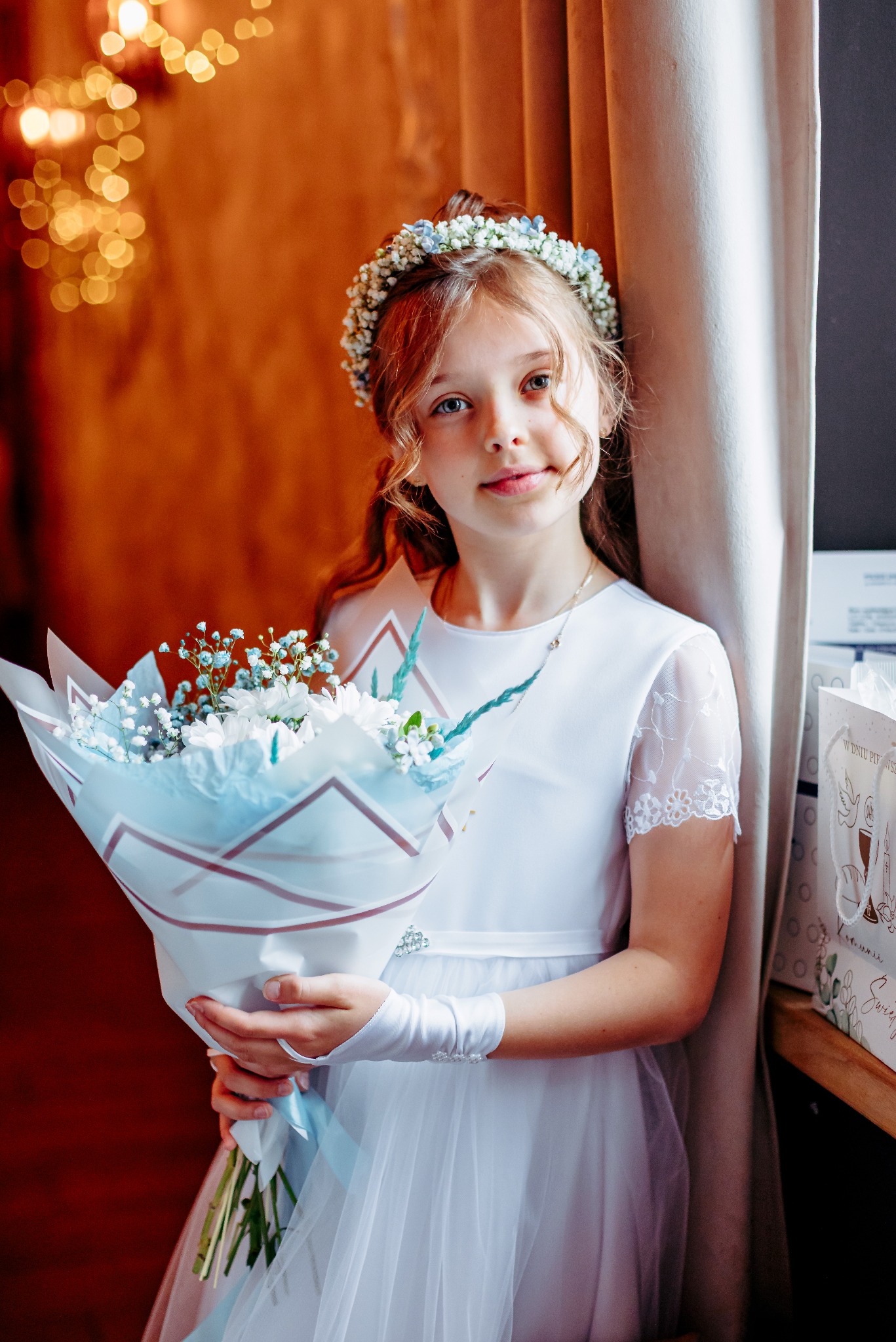Dziewczynka w białej sukience komunijnej, z wiankiem na głowie, trzyma bukiet białych kwiatów na tle okna i dekoracyjnej ściany. Delikatne światło.