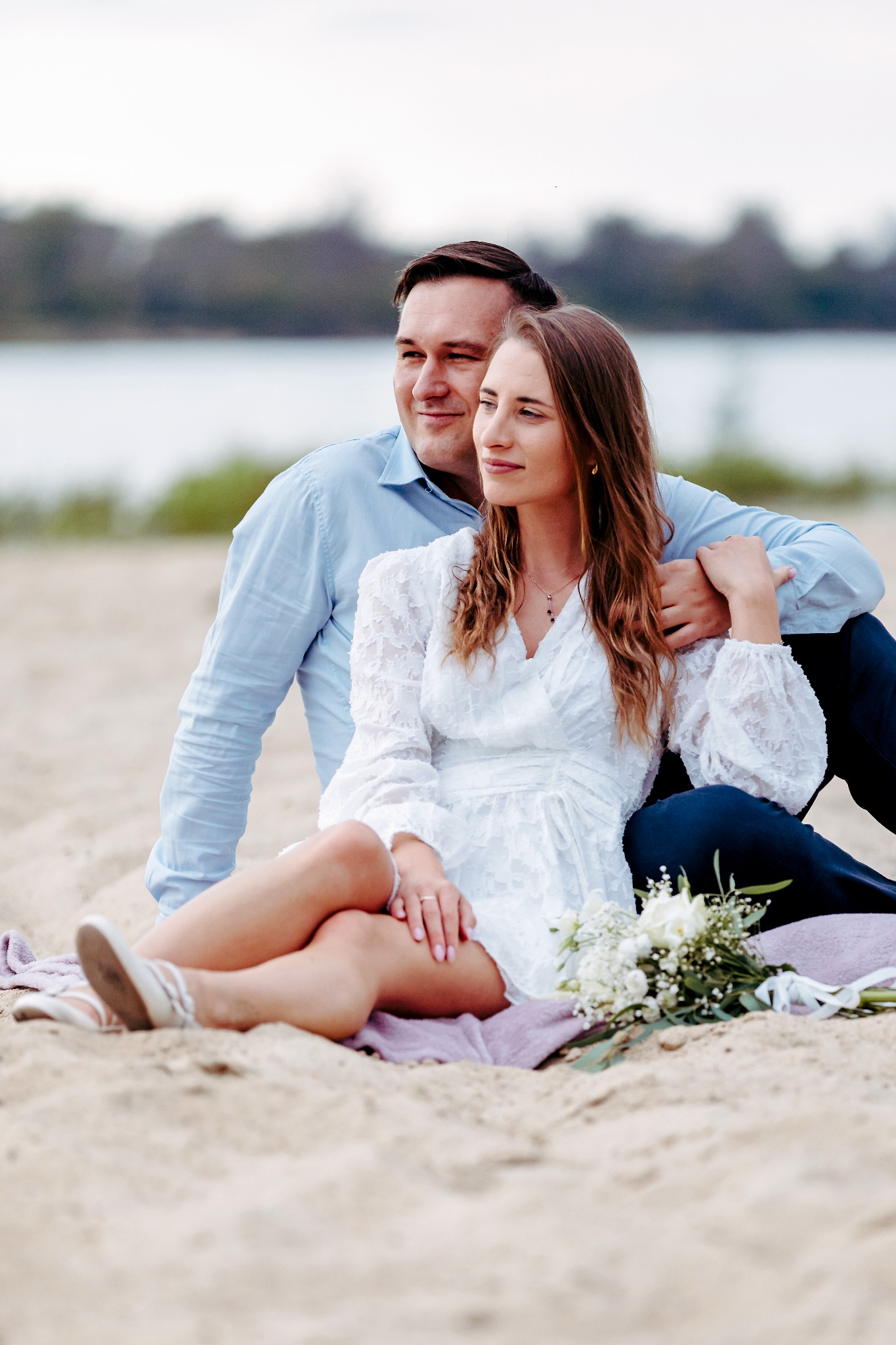 Para siedzi na piasku z bukietem kwiatów. Kobieta w białej sukience, mężczyzna w błękitnej koszuli. W tle woda i zieleń. Romantyczna sesja na plaży w naturalnym świetle.