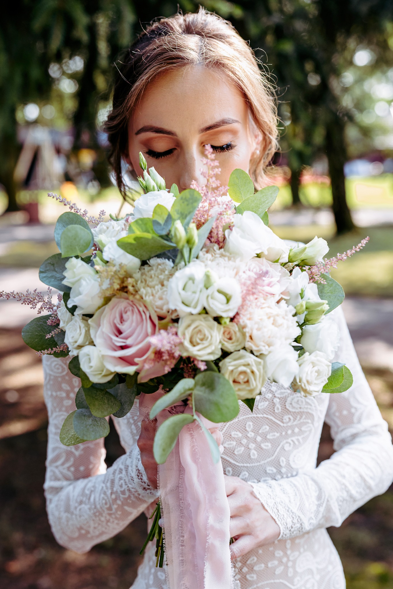 Panna młoda w koronkowej sukni, z zamkniętymi oczami, wącha bukiet ślubny z różowych i białych kwiatów, przewiązany różową wstążką. Delikatne światło podkreśla detale.
