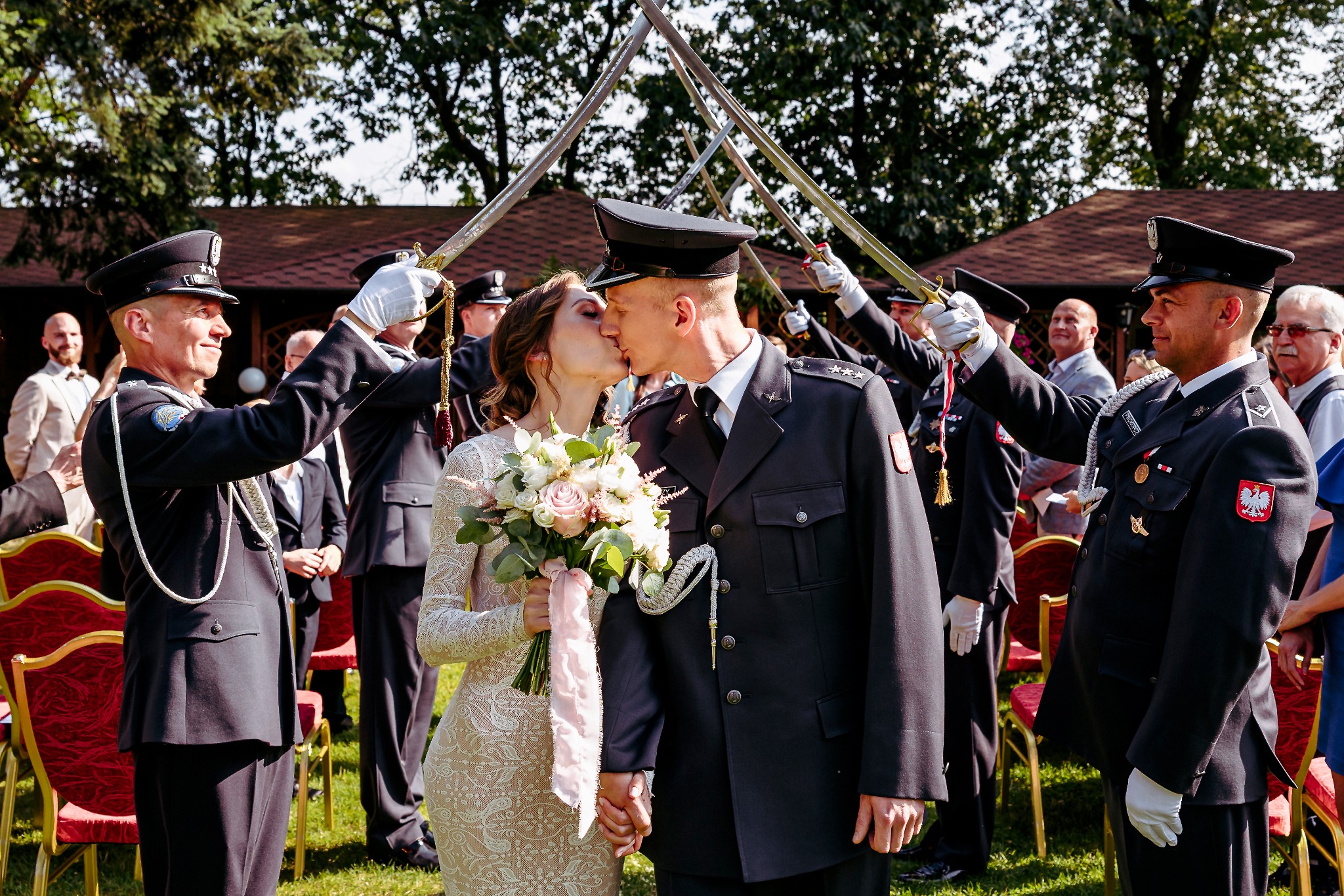 Pocałunek pary młodej w mundurach pod szpalerem szabel. Panna młoda w koronkowej sukni trzyma bukiet. Uroczystość na zewnątrz, w tle drzewa i czerwone krzesła.