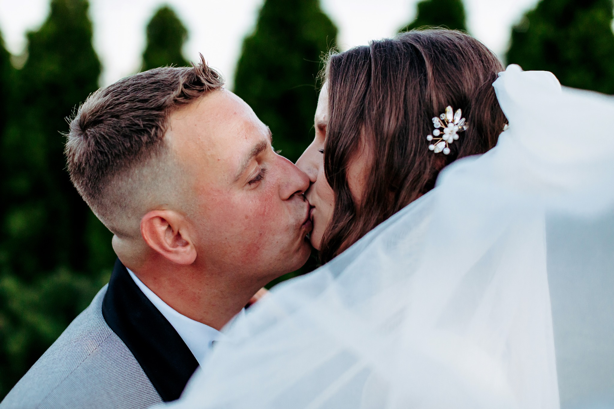Nowożeńcy w pocałunku: on w szarym smokingu, ona z ozdobą we włosach i welonem. Ujęcie z bliska, na zielonym tle. Romantyczna fotografia ślubna.