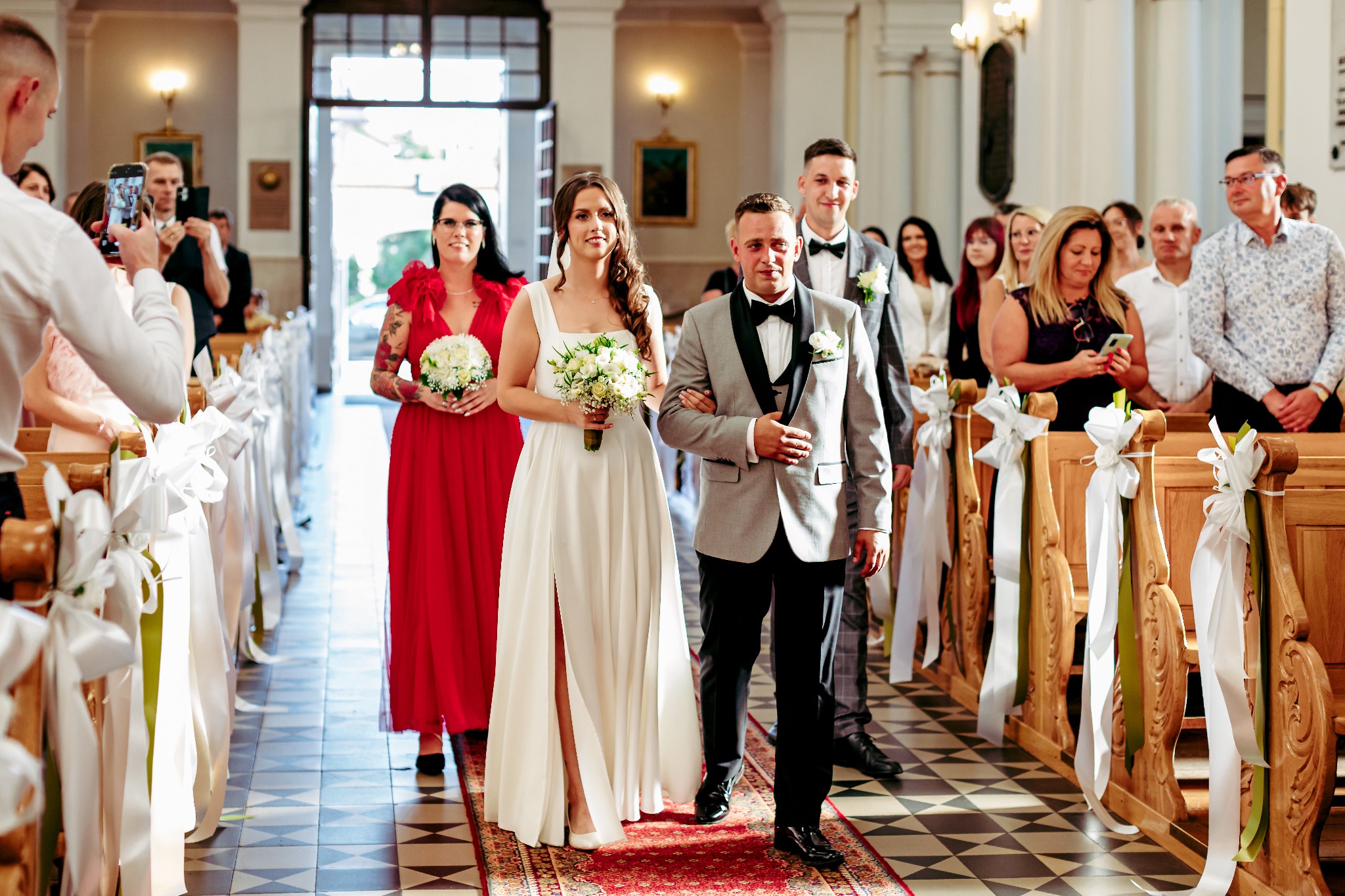 Para młoda prowadzona do ołtarza w kościele, goście obserwują i robią zdjęcia. Dekoracje z białych wstążek na ławkach. Panna młoda w sukni z rozcięciem.
