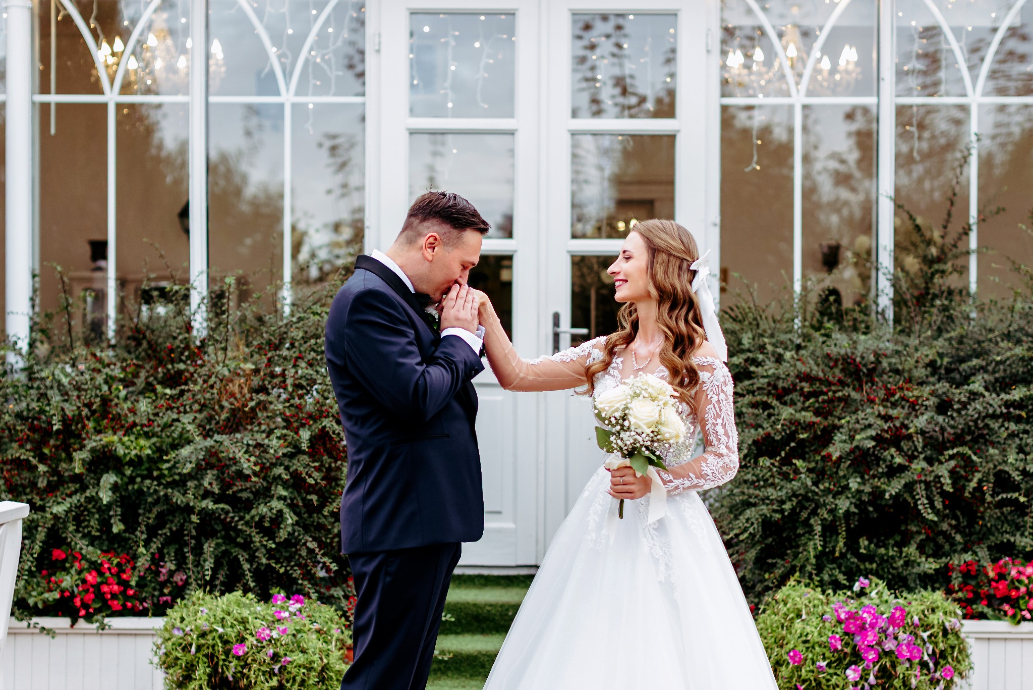 Elegancka para młoda: pan młody całuje dłoń uśmiechniętej panny młodej z bukietem białych róż na tle białych drzwi i zieleni.