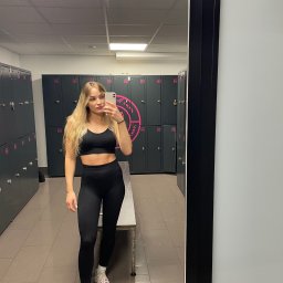 Barbara Obyrtacz - Blondwłosa kobieta w czarnym sportowym stroju robi selfie w szatni na tle szafek z numerami i logo siłowni. Widoczne odbicie w lustrze.