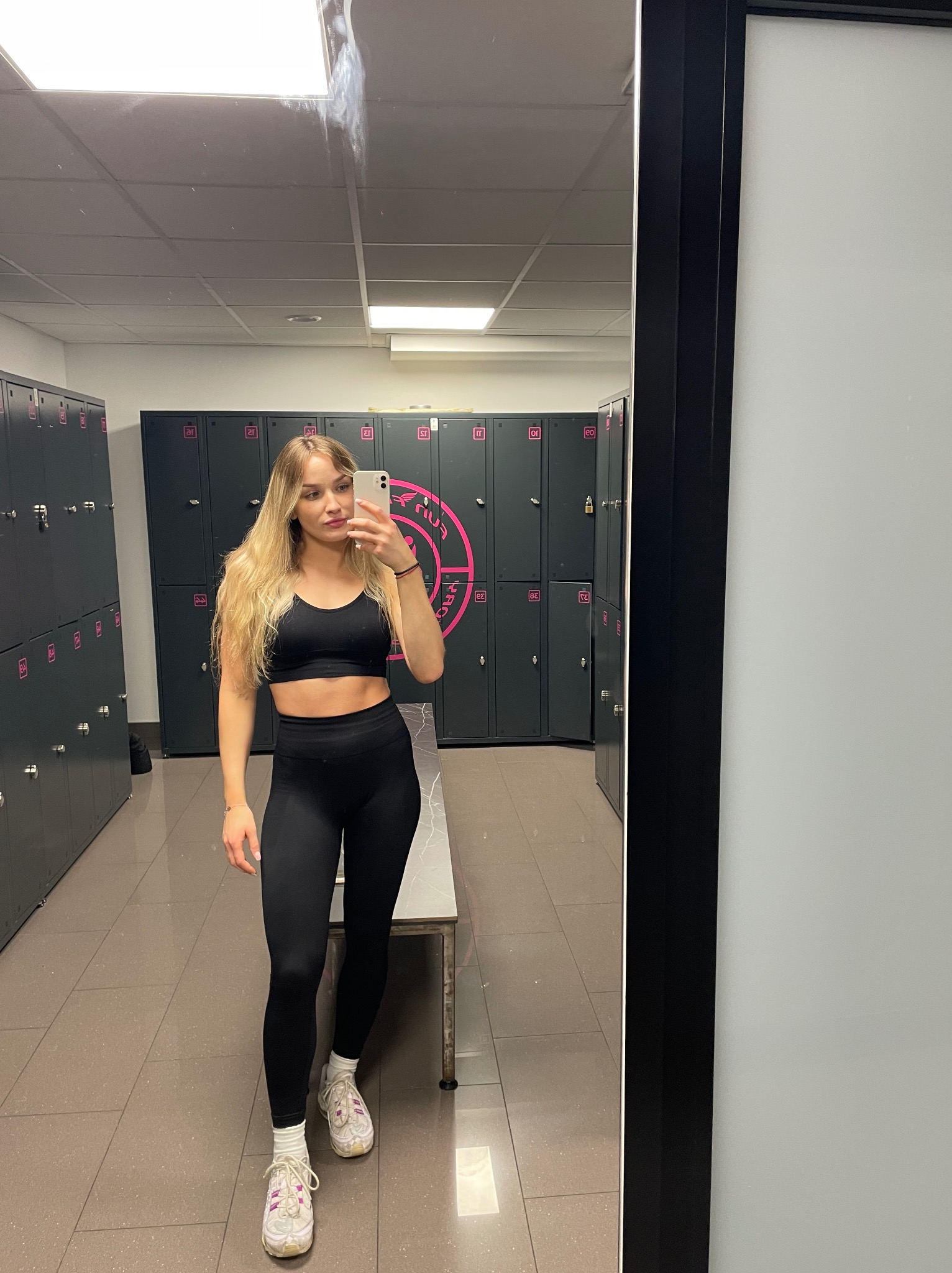 Blondwłosa kobieta w czarnym sportowym stroju robi selfie w szatni na tle szafek z numerami i logo siłowni. Widoczne odbicie w lustrze.