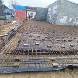 Usługi ogólno budowlane Dawid Mastalerz - Zbrojenie fundamentów na folii hydroizolacyjnej, widok z góry. W tle budynek w trakcie budowy i sprzęt budowlany. Prace zbrojarskie w toku.