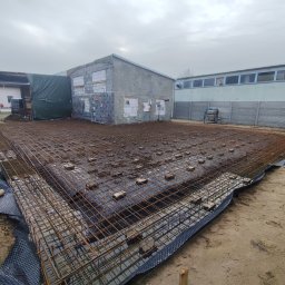 Usługi ogólno budowlane Dawid Mastalerz - Siatka zbrojeniowa na folii fundamentowej, przygotowana do zalania betonem. W tle budynek w trakcie budowy i pochmurne niebo. Widok z góry.