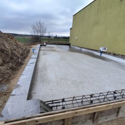 Usługi ogólno budowlane Dawid Mastalerz - Wylana płyta fundamentowa z widocznym zbrojeniem i szalunkiem drewnianym. Obok hałda ziemi i fragment budynku z żółtą elewacją. Pochmurne niebo w tle.