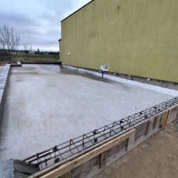 Usługi ogólno budowlane Dawid Mastalerz - Wylana płyta fundamentowa z szalunkiem i zbrojeniem, widok z góry. Świeży beton, szare bloczki fundamentowe, żółta ściana budynku w tle. Pochmurny dzień.