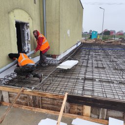 Usługi ogólno budowlane Dawid Mastalerz - Dwóch robotników w kamizelkach montuje rurę przy budynku. Widoczna siatka zbrojeniowa, szalunek i styropian. Prace przygotowawcze do wylewki fundamentów.