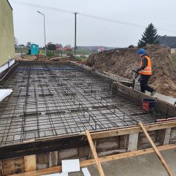 Usługi ogólno budowlane Dawid Mastalerz - Zbrojenie fundamentów pod budynek jednorodzinny, widoczna siatka zbrojeniowa, szalunek drewniany i pracownik w odblaskowej kamizelce. Prace budowlane w toku.