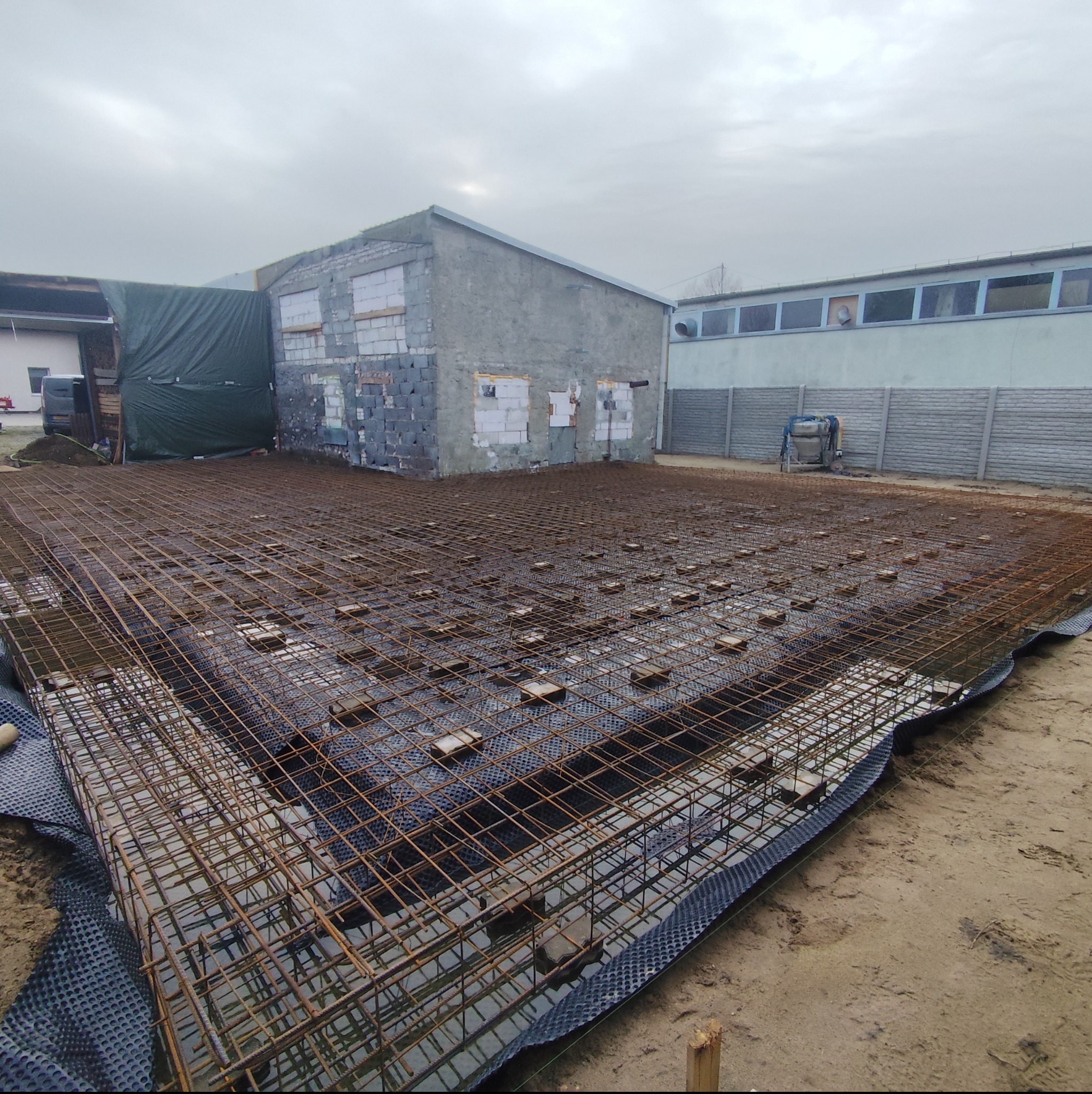 Siatka zbrojeniowa na folii fundamentowej, przygotowana do zalania betonem. W tle budynek w trakcie budowy i pochmurne niebo. Widok z góry.