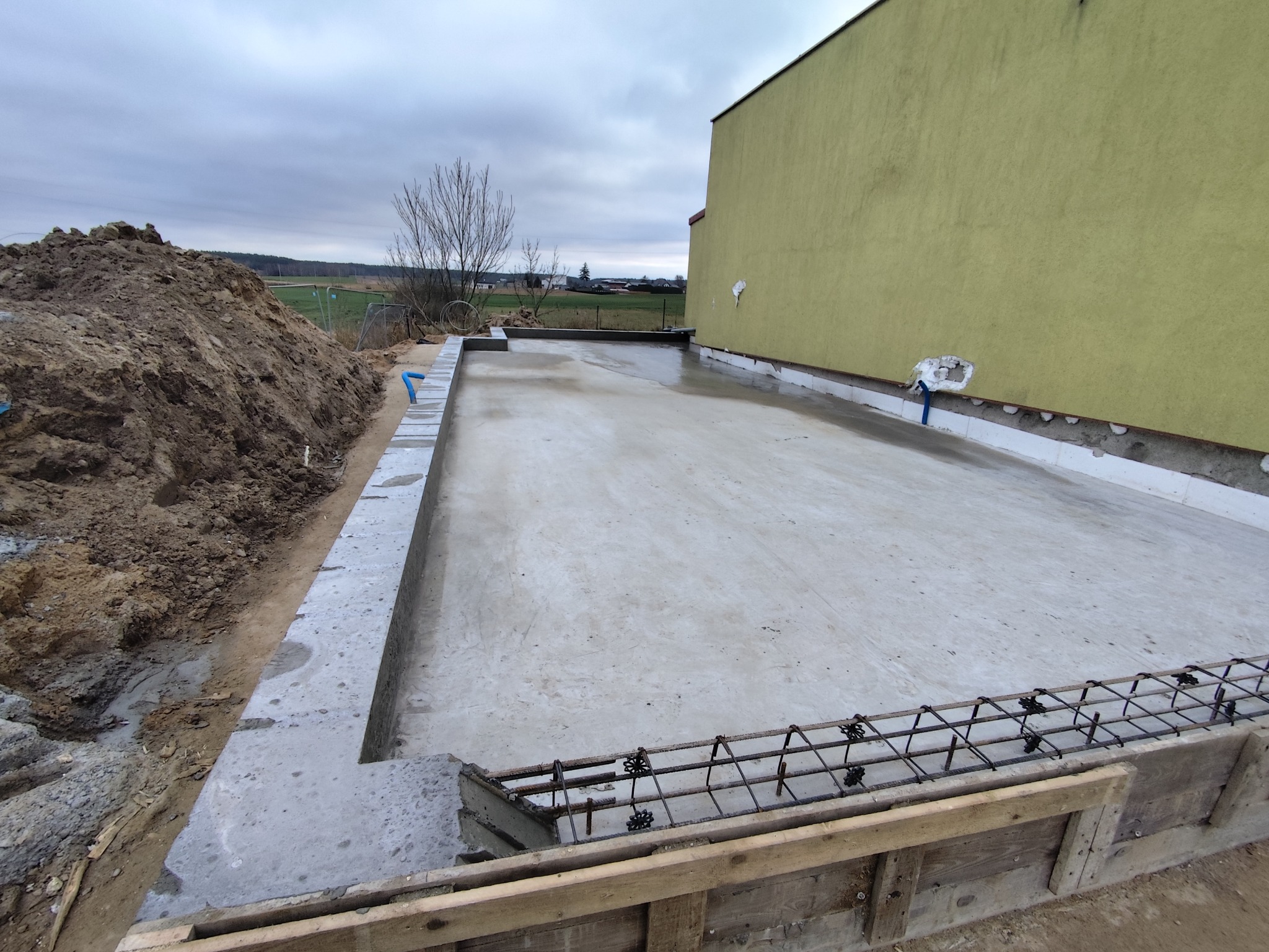 Wylana płyta fundamentowa z widocznym zbrojeniem i szalunkiem drewnianym. Obok hałda ziemi i fragment budynku z żółtą elewacją. Pochmurne niebo w tle.