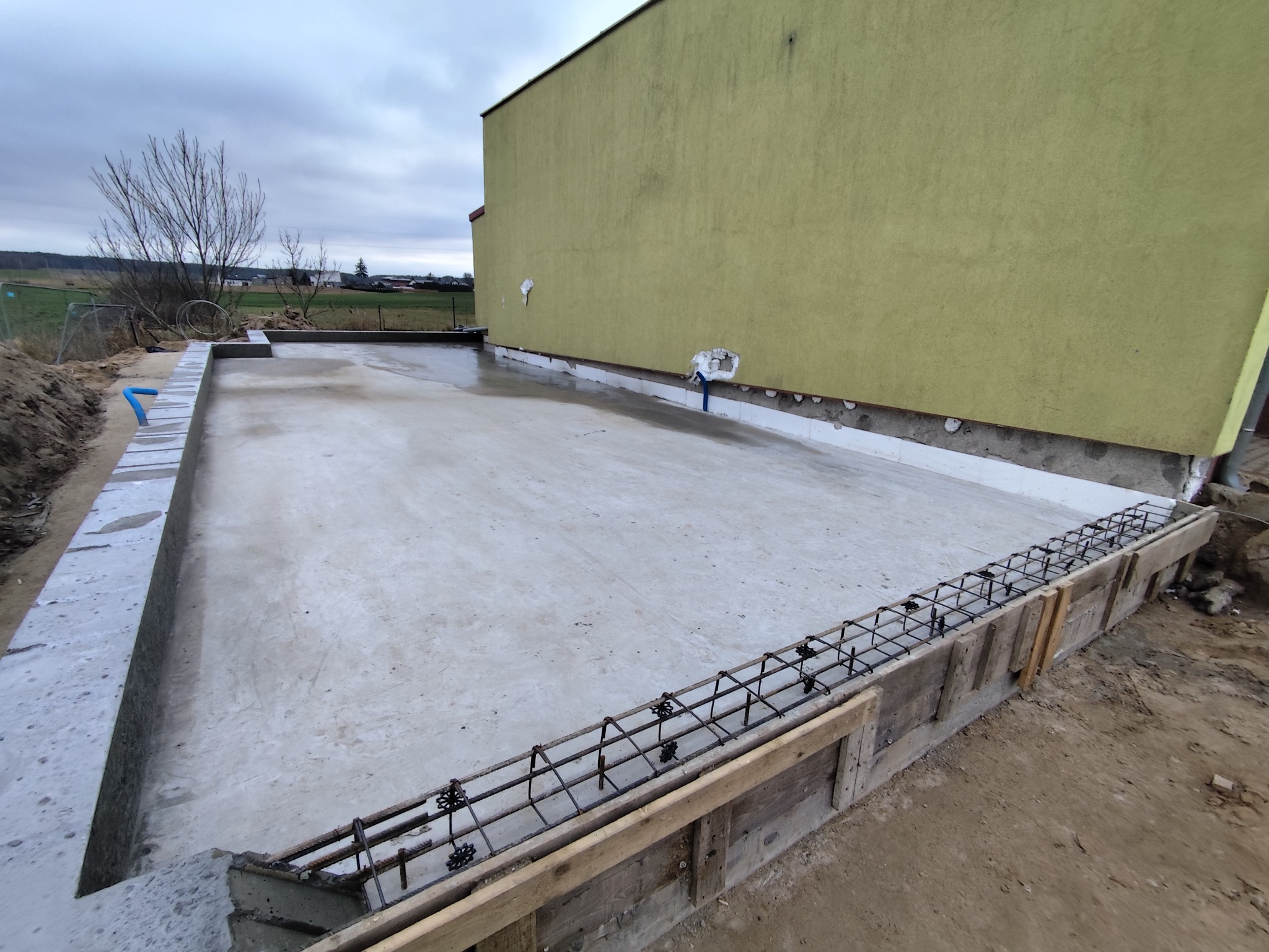Wylana płyta fundamentowa z szalunkiem i zbrojeniem, widok z góry. Świeży beton, szare bloczki fundamentowe, żółta ściana budynku w tle. Pochmurny dzień.