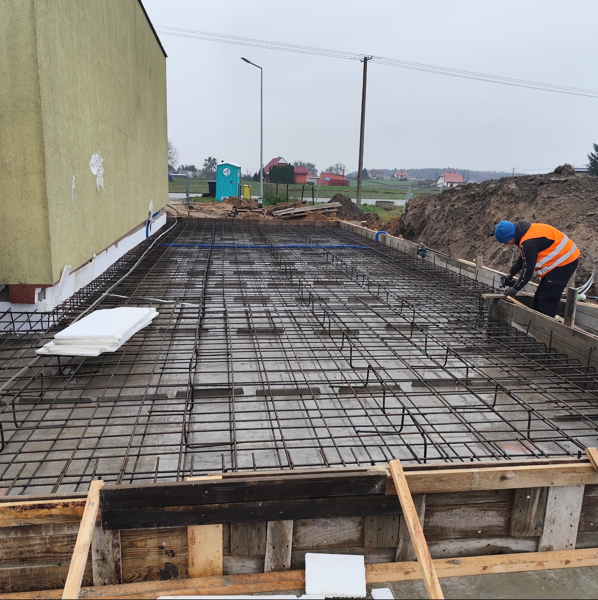Zbrojenie płyty fundamentowej z deskowaniem i pracującym zbrojarzem w pomarańczowej kamizelce. Widoczne białe płyty izolacyjne i niebieskie rurki instalacyjne.