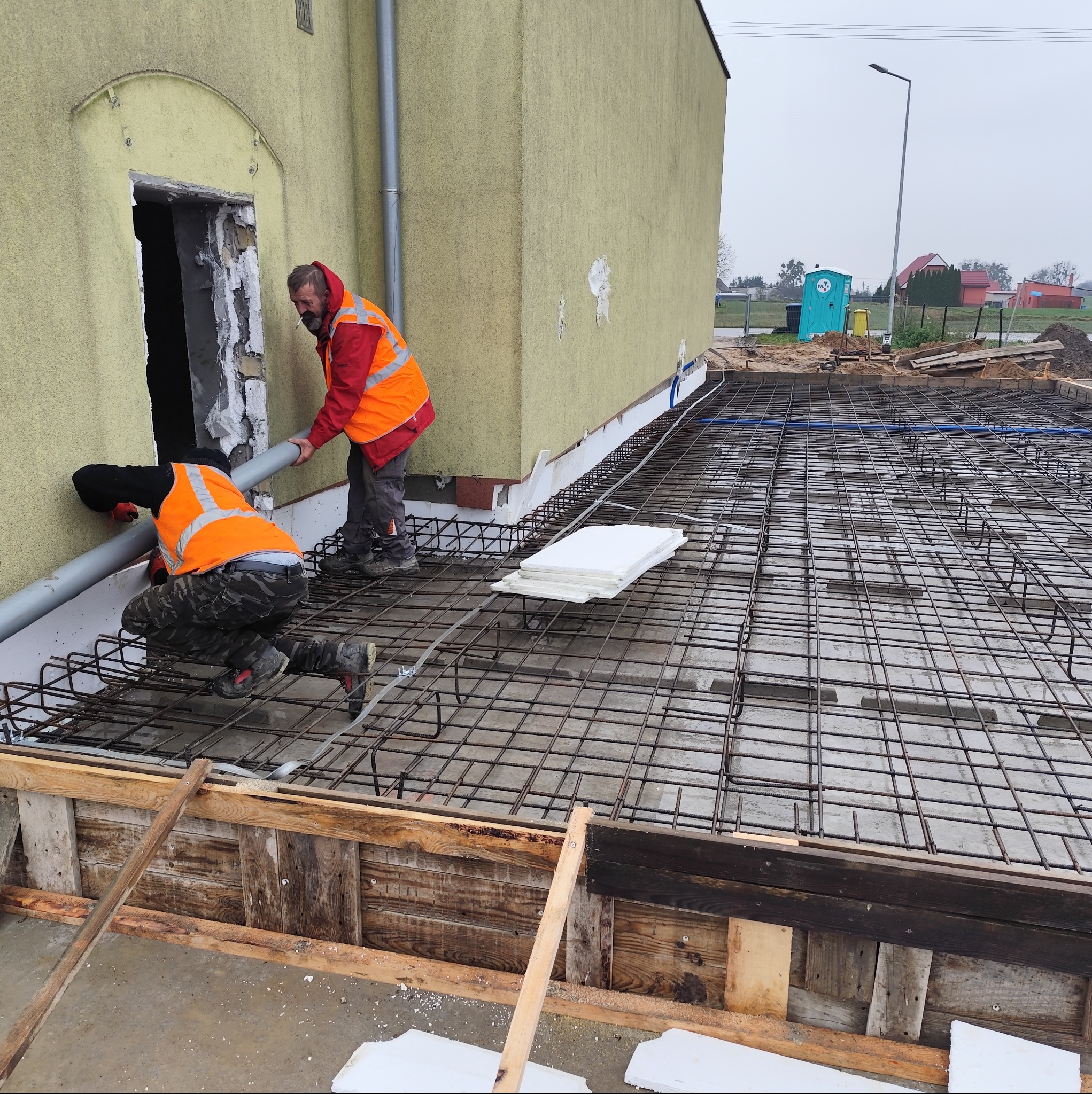 Dwóch robotników w kamizelkach montuje rurę przy budynku. Widoczna siatka zbrojeniowa, szalunek i styropian. Prace przygotowawcze do wylewki fundamentów.