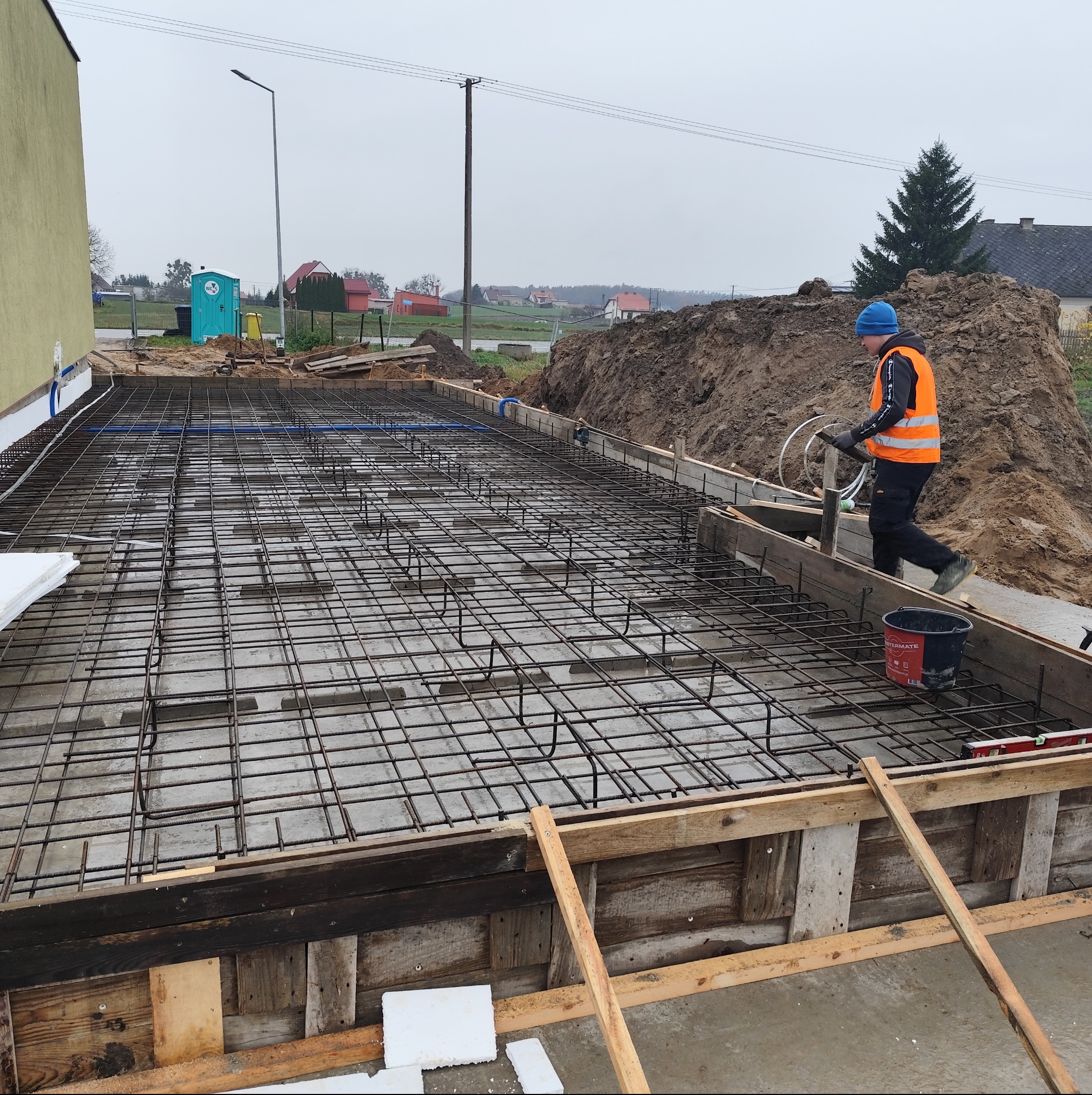 Zbrojenie fundamentów pod budynek jednorodzinny, widoczna siatka zbrojeniowa, szalunek drewniany i pracownik w odblaskowej kamizelce. Prace budowlane w toku.
