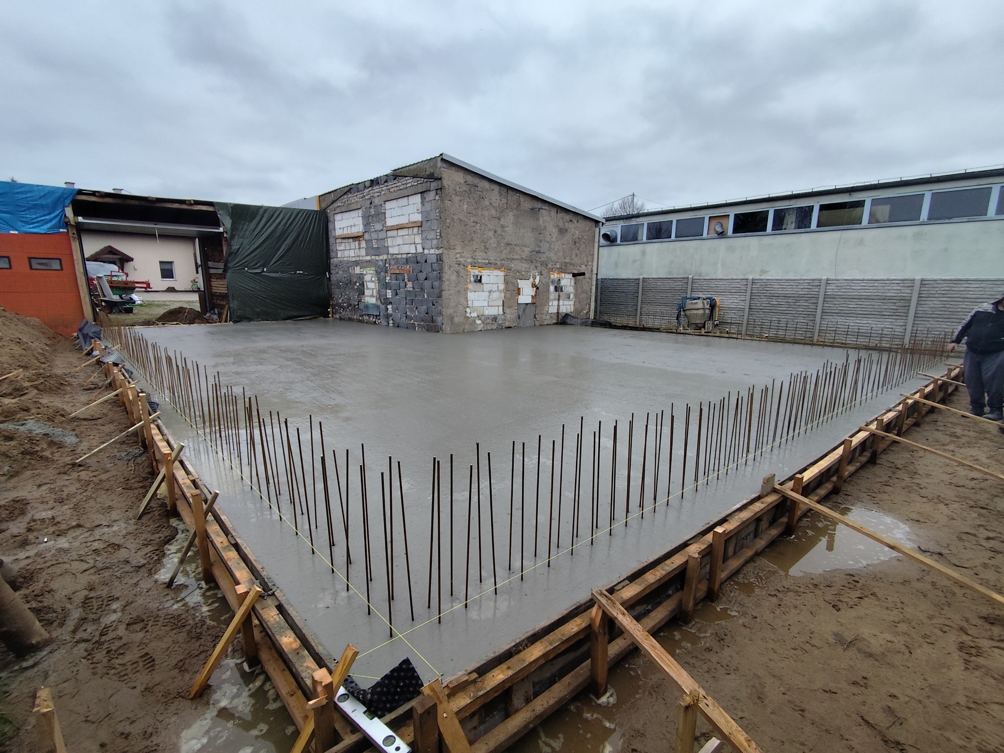 Zalany betonem fundament z wystającymi prętami zbrojeniowymi, drewnianym szalunkiem i budynkiem w tle, w pochmurny dzień. Widok z poziomu gruntu.