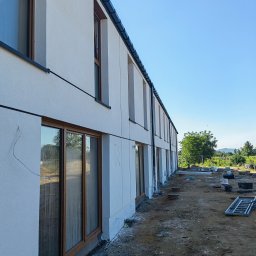 GIFTPOL Paweł Gołubców - Biała elewacja budynku z drewnianymi oknami, widok z boku, teren budowy w tle. Minimalistyczny styl, jasne oświetlenie, błękitne niebo.