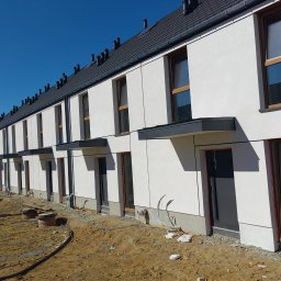 GIFTPOL Paweł Gołubców - Rząd nowych, dwukondygnacyjnych domów szeregowych z białymi elewacjami i ciemnymi dachami, w trakcie wykończenia terenu wokół budynków. Widok z zewnątrz.