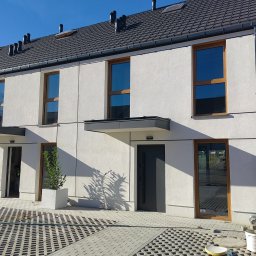 GIFTPOL Paweł Gołubców - Nowoczesny segment domu w zabudowie szeregowej z jasną elewacją o fakturze baranka, drewnianą stolarką okienną i ciemną dachówką. Widoczne detale wykończenia.