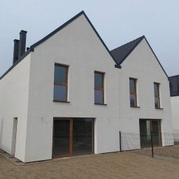 GIFTPOL Paweł Gołubców - Nowoczesny dom z białej cegły z ciemną dachówką i stolarką okienną. Minimalistyczny design, ogrodzenie panelowe. Widok z zewnątrz, w tle drugi budynek.