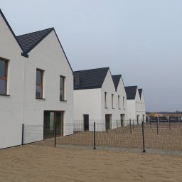 GIFTPOL Paweł Gołubców - Szereg nowych, białych domów z ciemnymi dachami, ogrodzonych siatką. Widok z zewnątrz, w tle krajobraz. Minimalistyczna architektura, równomierne oświetlenie.