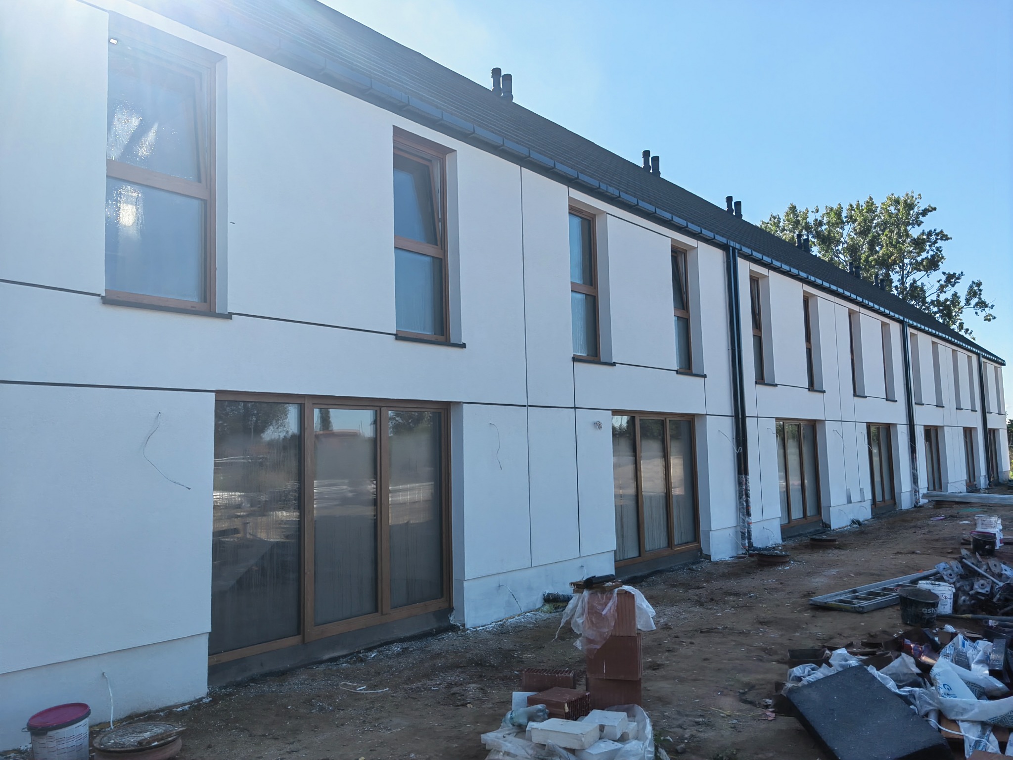 Nowoczesny budynek w trakcie wykończenia elewacji, z widocznymi oknami z drewnianymi ramami i elementami konstrukcyjnymi. Na pierwszym planie teren budowy.