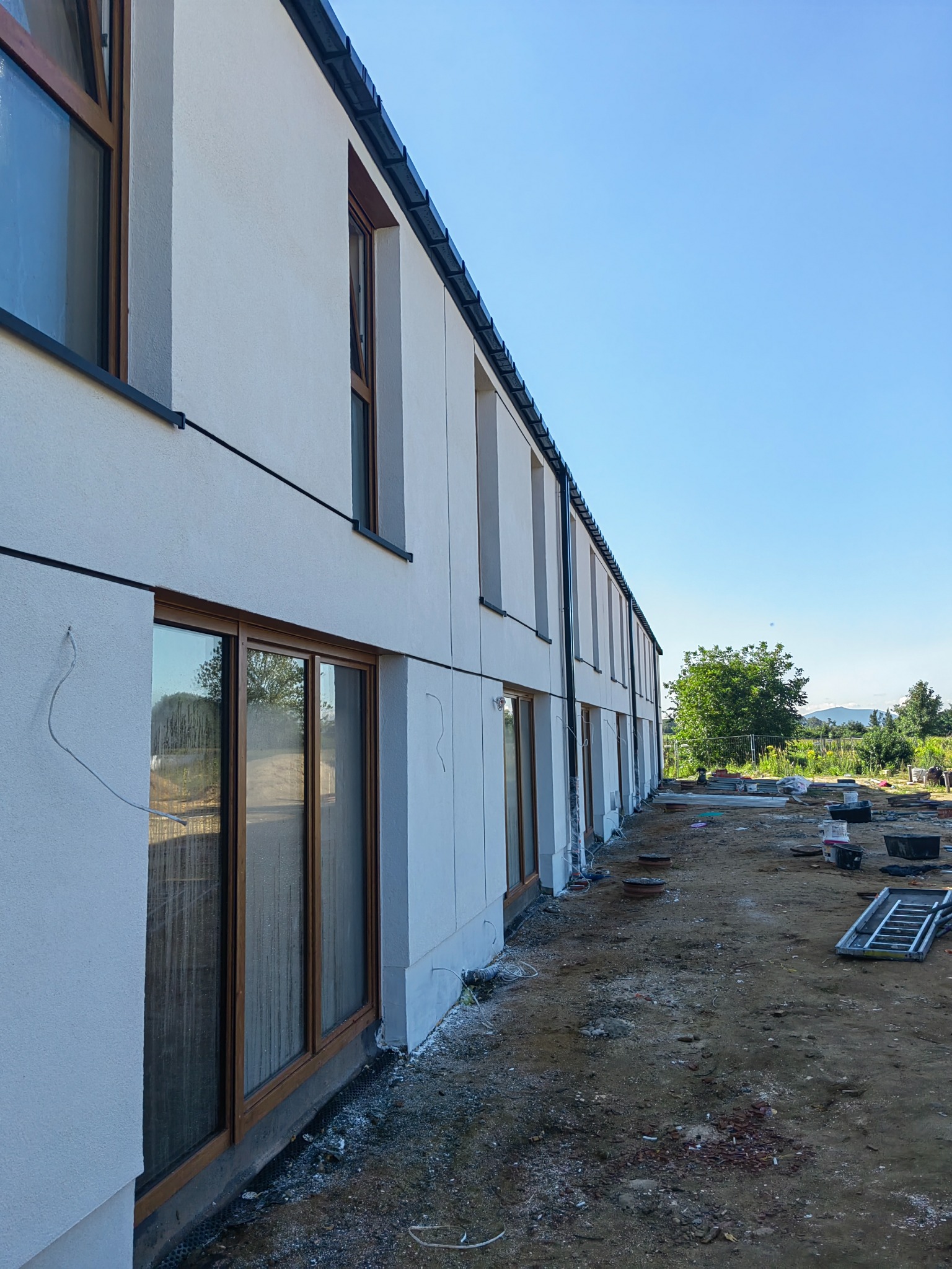 Biała elewacja budynku z drewnianymi oknami, widok z boku, teren budowy w tle. Minimalistyczny styl, jasne oświetlenie, błękitne niebo.