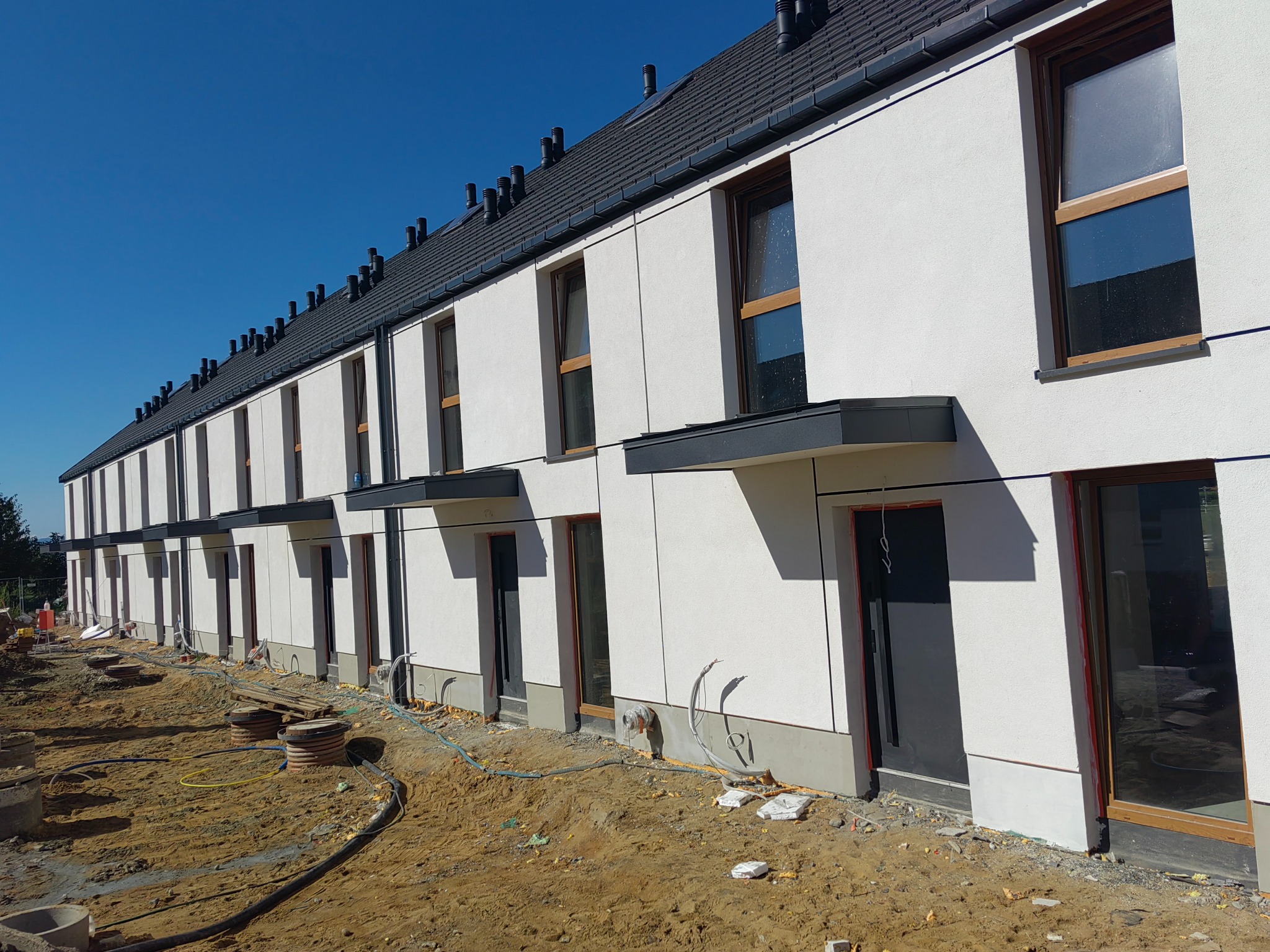 Rząd nowych, dwukondygnacyjnych domów szeregowych z białymi elewacjami i ciemnymi dachami, w trakcie wykończenia terenu wokół budynków. Widok z zewnątrz.