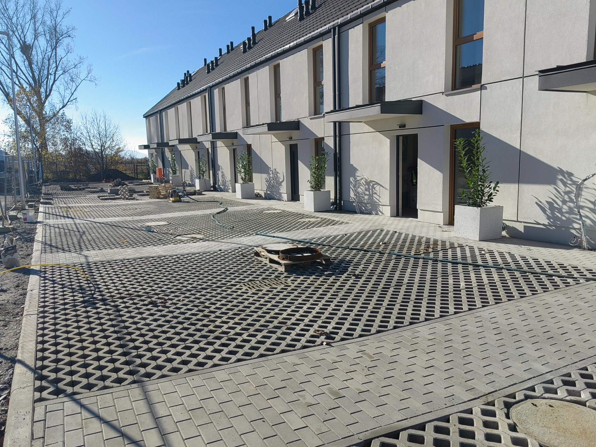 Nowoczesne domy szeregowe z jasną elewacją i ażurowym parkingiem z kostki. Widoczne rośliny w donicach przy wejściach. Słoneczny dzień, błękitne niebo.