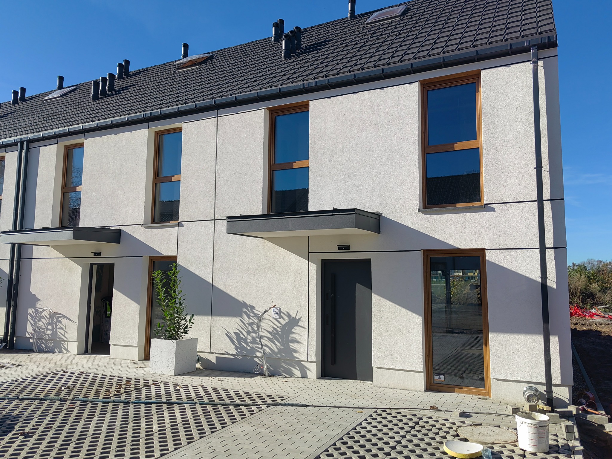 Nowoczesny segment domu w zabudowie szeregowej z jasną elewacją o fakturze baranka, drewnianą stolarką okienną i ciemną dachówką. Widoczne detale wykończenia.