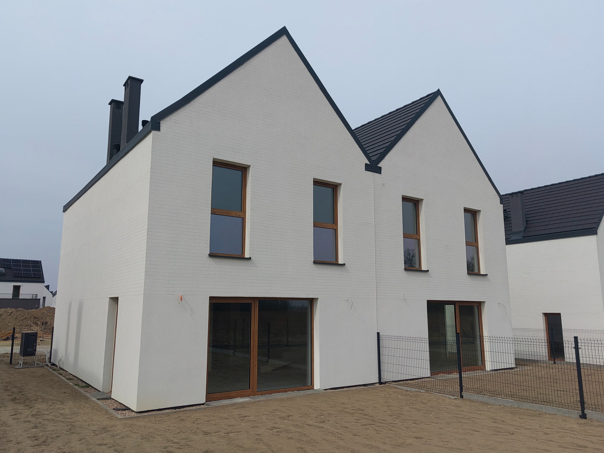 Nowoczesny dom z białej cegły z ciemną dachówką i stolarką okienną. Minimalistyczny design, ogrodzenie panelowe. Widok z zewnątrz, w tle drugi budynek.