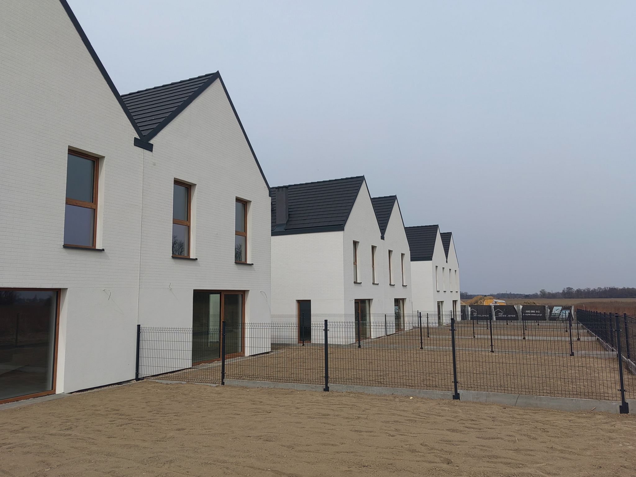 Szereg nowych, białych domów z ciemnymi dachami, ogrodzonych siatką. Widok z zewnątrz, w tle krajobraz. Minimalistyczna architektura, równomierne oświetlenie.