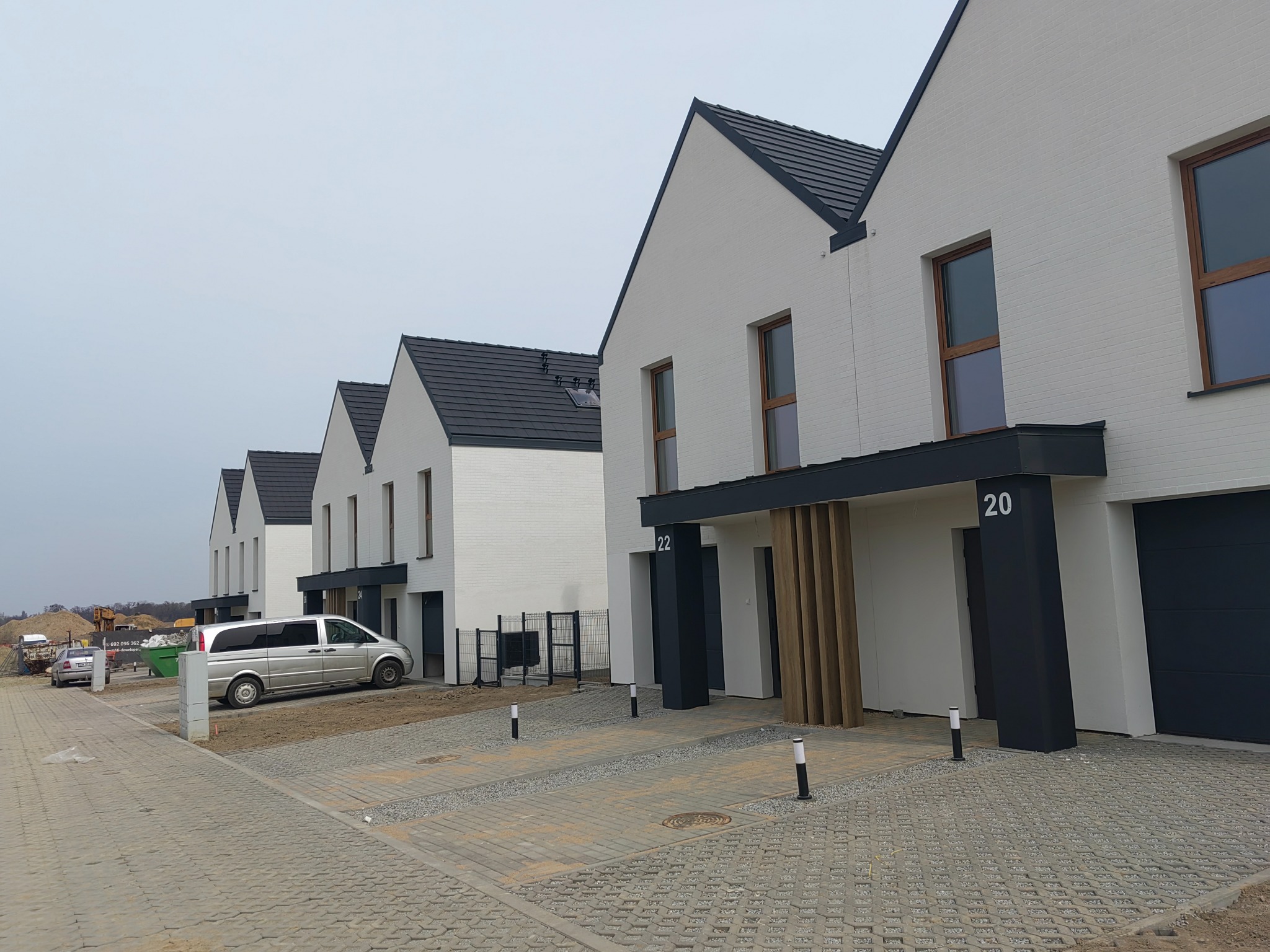 Szereg identycznych, nowoczesnych domów z białymi elewacjami i ciemnymi dachami, z numerami 20 i 22, widzianych z perspektywy ulicy, w tle plac budowy.