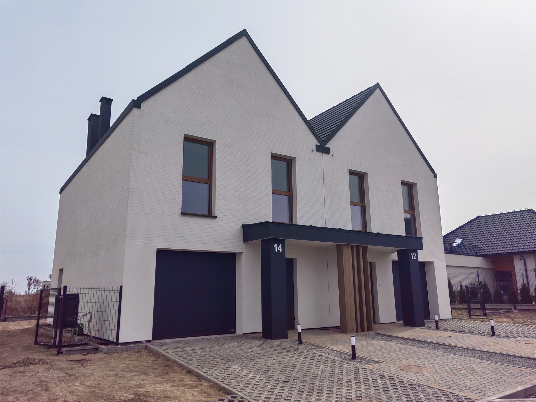 Biały, dwukondygnacyjny dom bliźniak z ciemną stolarką okienną i garażem. Elewacja z cegły klinkierowej, czarny dach i numeracja budynków na kolumnach wejściowych.