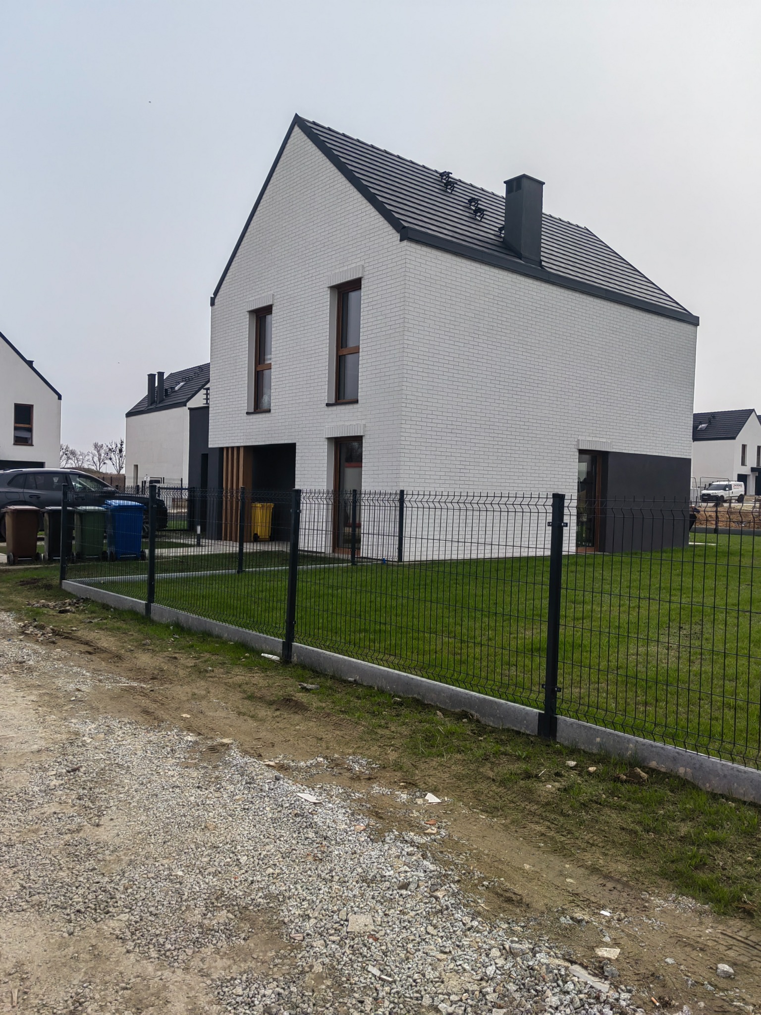 Nowoczesny dom z białej cegły z ciemną stolarką okienną i dachem, otoczony zielonym trawnikiem i ogrodzeniem z siatki. Widok z poziomu gruntu na osiedlu.