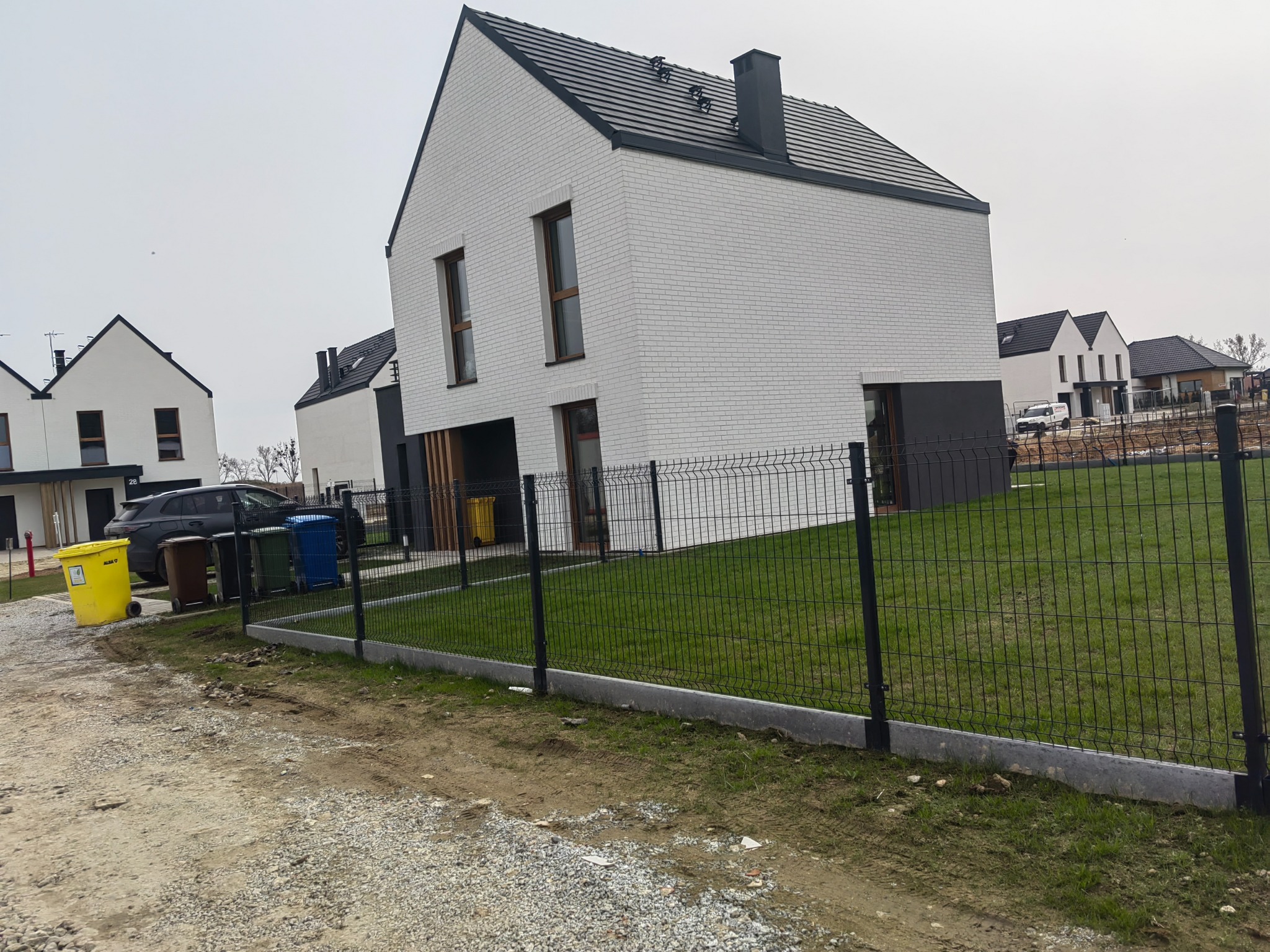 Nowoczesny dom z białej cegły z ciemną stolarką i dachem, ogrodzony metalową siatką. Widoczne inne domy w tle, pochmurne niebo.