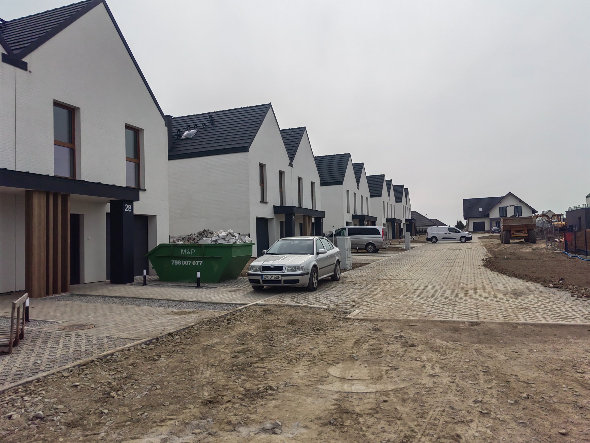 Rząd nowych, białych domów z ciemnymi dachami w trakcie budowy. Widoczny kontener z gruzem, samochód osobowy i teren z kostką brukową oraz ziemią.