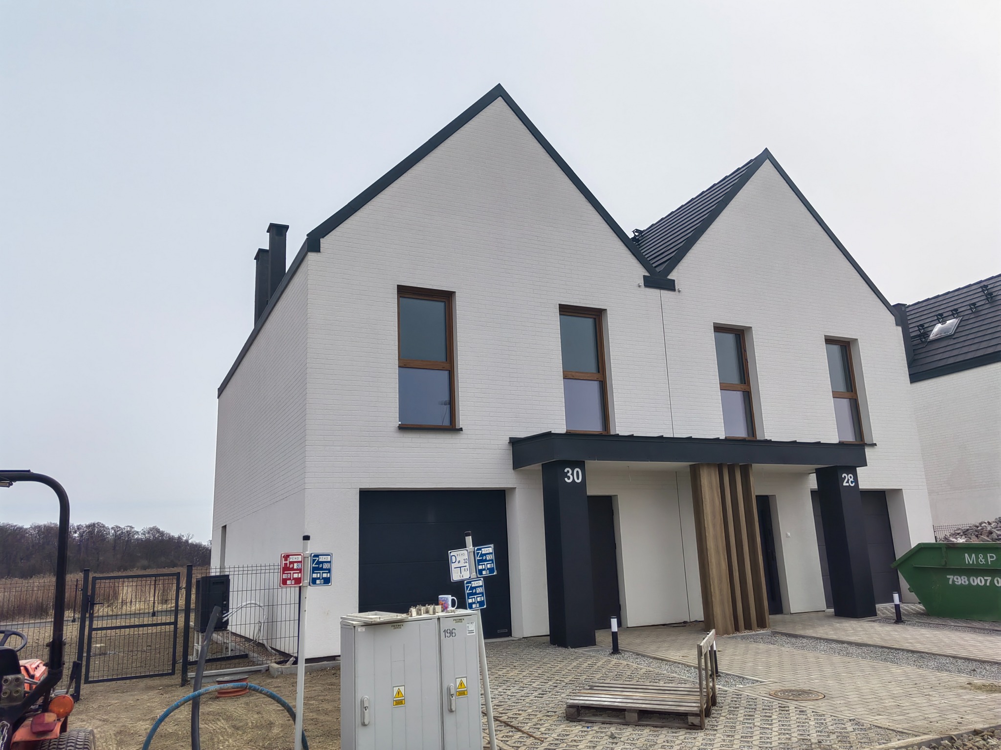 Nowoczesne domy bliźniacze z białej cegły, czarnymi dachami i drewnianymi akcentami. Widoczne numery domów 28 i 30. Teren wokół w trakcie zagospodarowania.