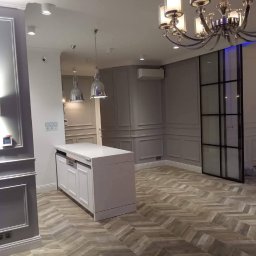 MG builders design company - Eleganckie wykończenie wnętrza z parkietem w jodełkę, sztukaterią na ścianach i nowoczesnym oświetleniem. Widoczna wyspa kuchenna i przeszklone drzwi.