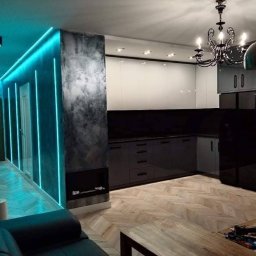 MG builders design company - Nowoczesne wnętrze: salon z aneksem kuchennym. Ściana z oświetleniem LED, parkiet w jodełkę, czarne meble kuchenne i dekoracyjny żyrandol. Widok na wykończone pomieszczenie.