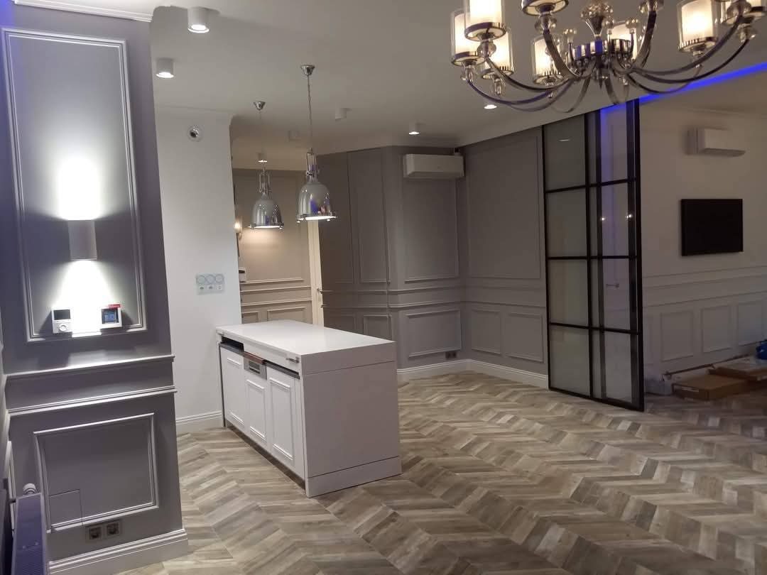 Eleganckie wykończenie wnętrza z parkietem w jodełkę, sztukaterią na ścianach i nowoczesnym oświetleniem. Widoczna wyspa kuchenna i przeszklone drzwi.