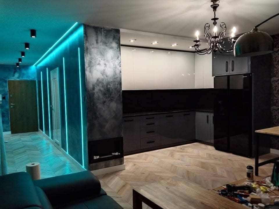 Nowoczesne wnętrze: salon z aneksem kuchennym. Ściana z oświetleniem LED, parkiet w jodełkę, czarne meble kuchenne i dekoracyjny żyrandol. Widok na wykończone pomieszczenie.