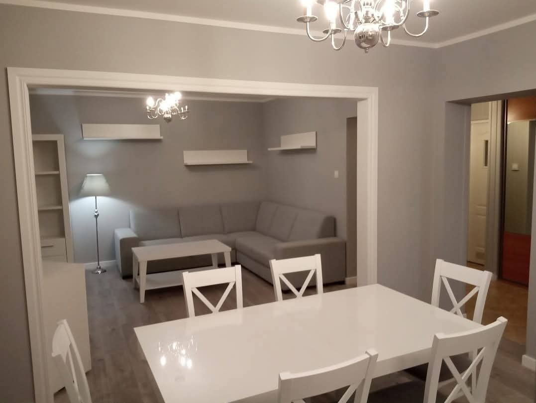 Jasne, szare wnętrze po remoncie: salon z narożnikiem i jadalnią. Białe meble, lampa podłogowa i żyrandol. Widok na pokój przez szerokie przejście.