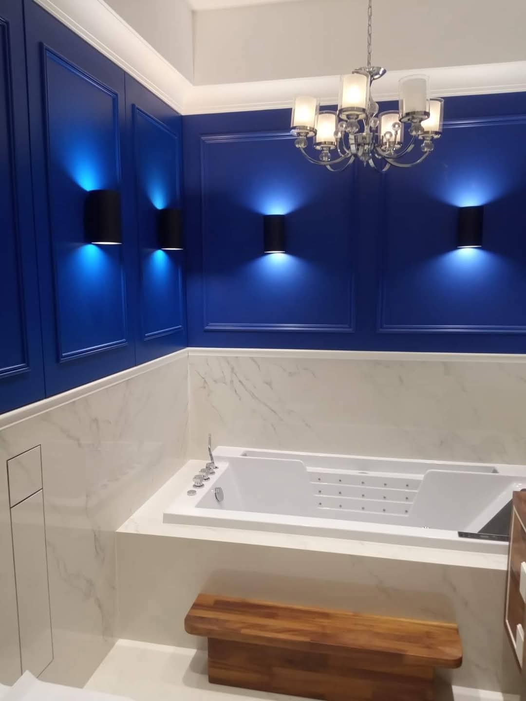 Niebieska łazienka z wanną jacuzzi, marmurowymi płytkami i designerskim oświetleniem: czarne kinkiety i kryształowy żyrandol. Drewniany stołek.