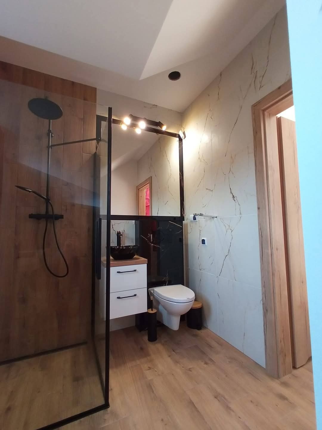 Nowoczesna łazienka z kabiną prysznicową typu walk-in, umywalką nablatową i toaletą. Drewnopodobne płytki i marmurowe akcenty tworzą elegancki wystrój.